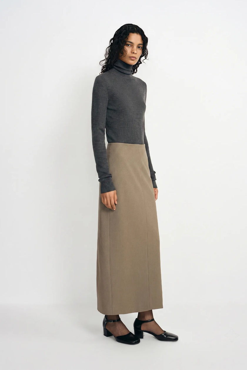 A model stands in a studio wearing a dark gray mock-neck long-sleeve top and a high-waisted, knee-length taupe skirt, paired with black shoes.