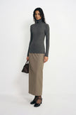 A model stands against a plain white studio background wearing a dark gray ribbed turtleneck sweater and a beige midi skirt, holding a dark brown handbag.