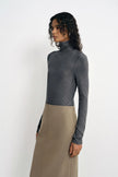 A model with curly dark hair stands in three-quarter profile against a plain white background, wearing a charcoal ribbed turtleneck top and a khaki/taupe midi skirt.