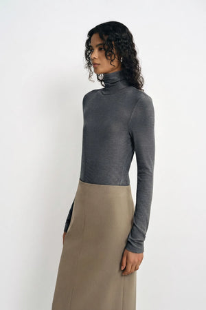 A model with curly dark hair stands in three-quarter profile against a plain white background, wearing a charcoal ribbed turtleneck top and a khaki/taupe midi skirt.