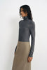 A model with curly dark hair stands in three-quarter profile against a plain white background, wearing a charcoal ribbed turtleneck top and a khaki/taupe midi skirt.
