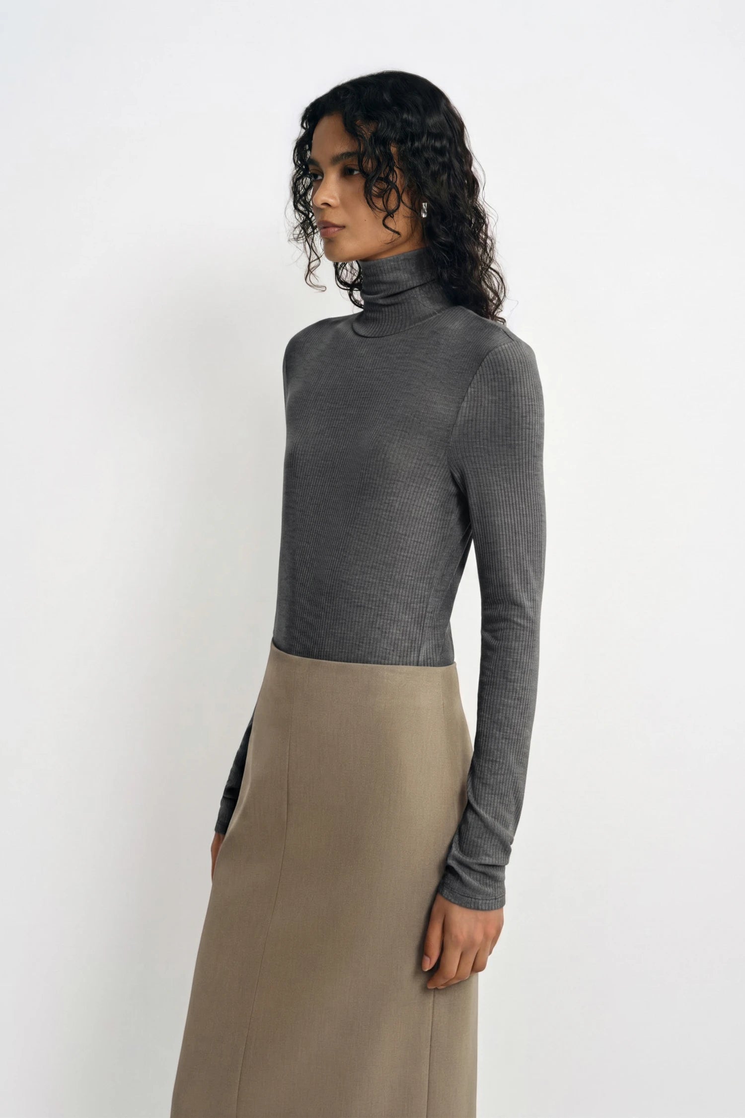 A model wearing a dark gray long-sleeve mock neck top paired with a beige midi skirt, photographed against a plain white studio background.
