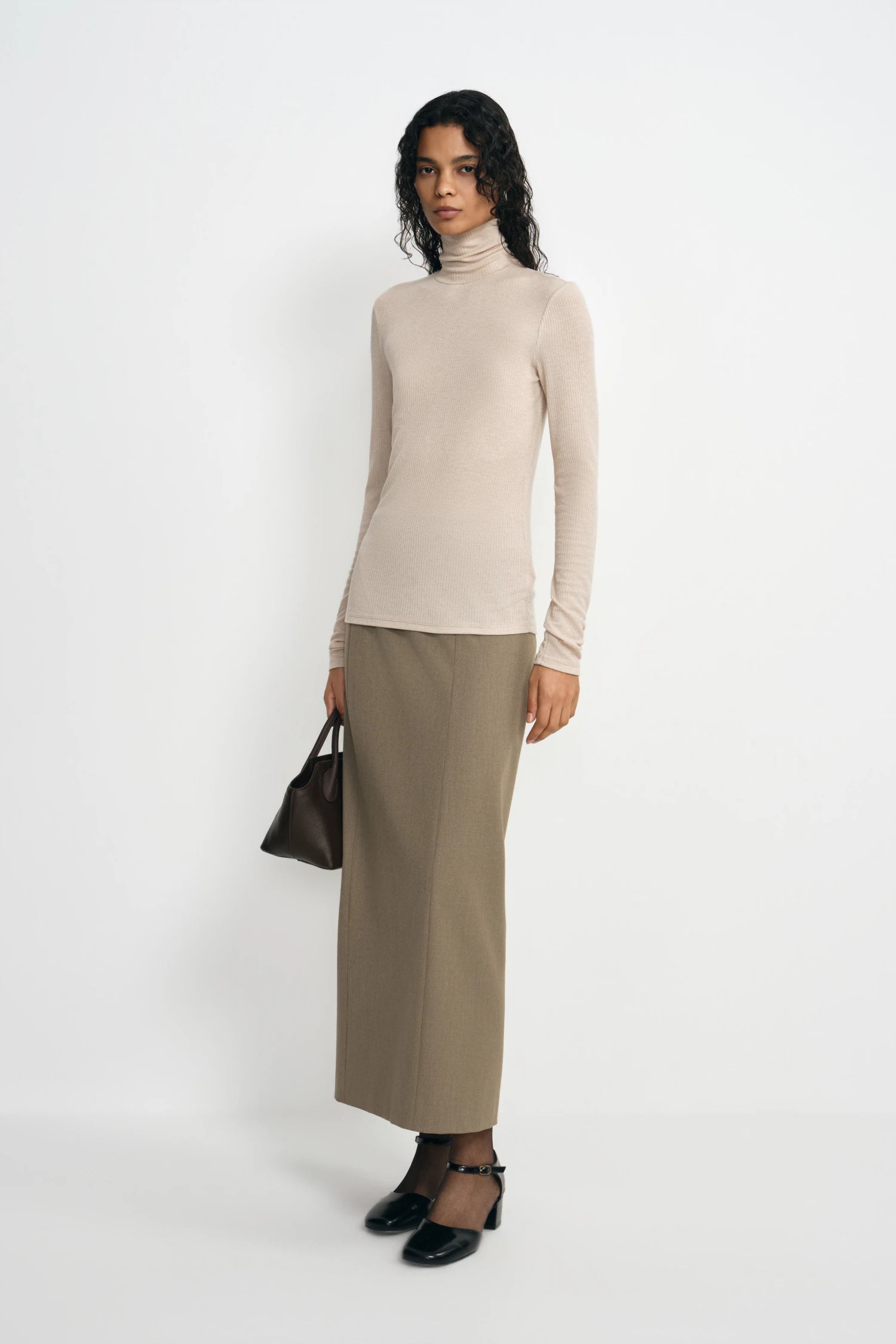 A model wearing a beige ribbed turtleneck top, pairing an oatmeal column skirt, turned slightly to the side.