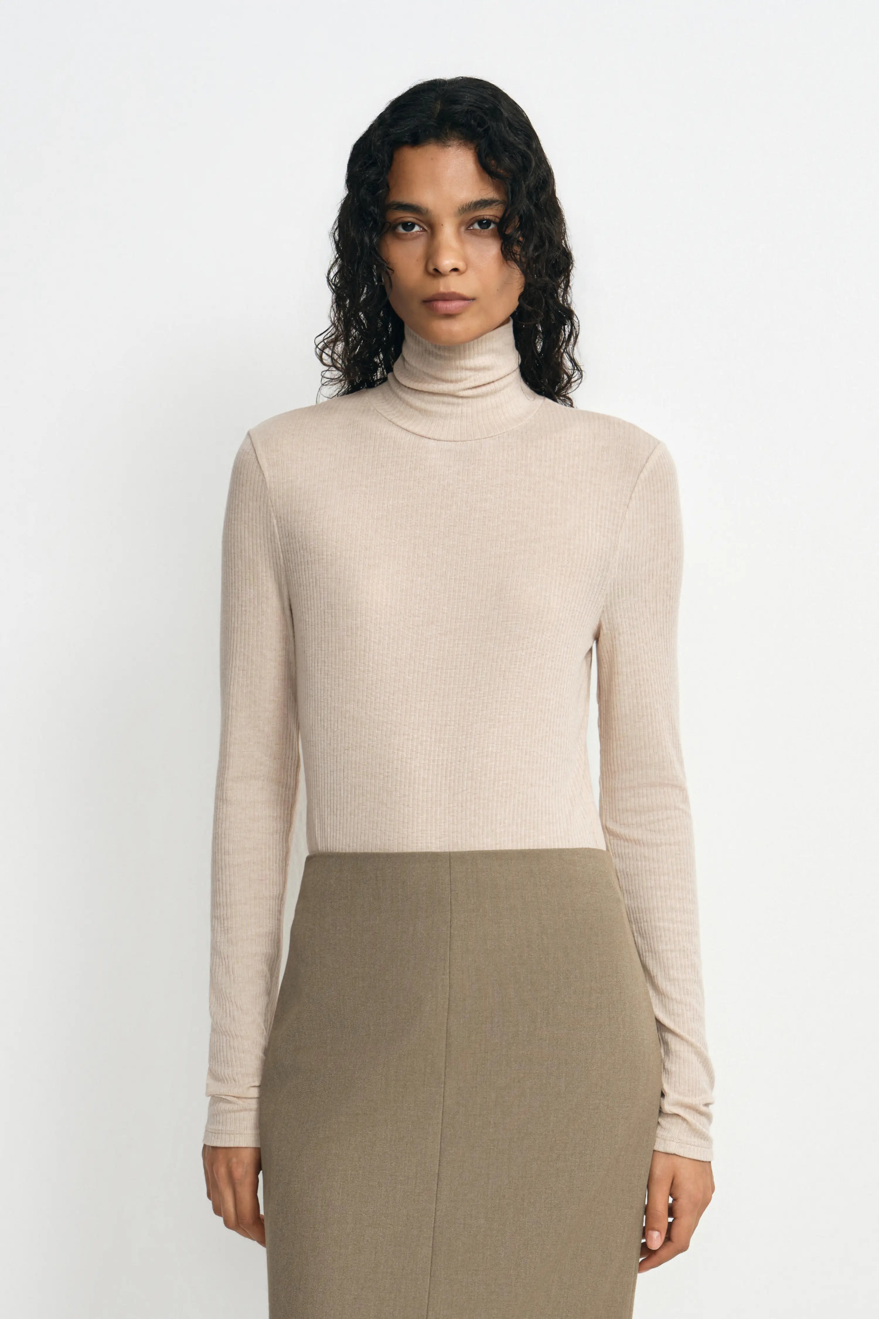 A model wearing a light beige ribbed turtleneck sweater and a taupe pencil skirt, standing against a plain white studio background.