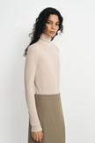 A woman modeling a ribbed long-sleeve turtleneck sweater in a light beige color, paired with a taupe skirt, against a plain light gray studio background.