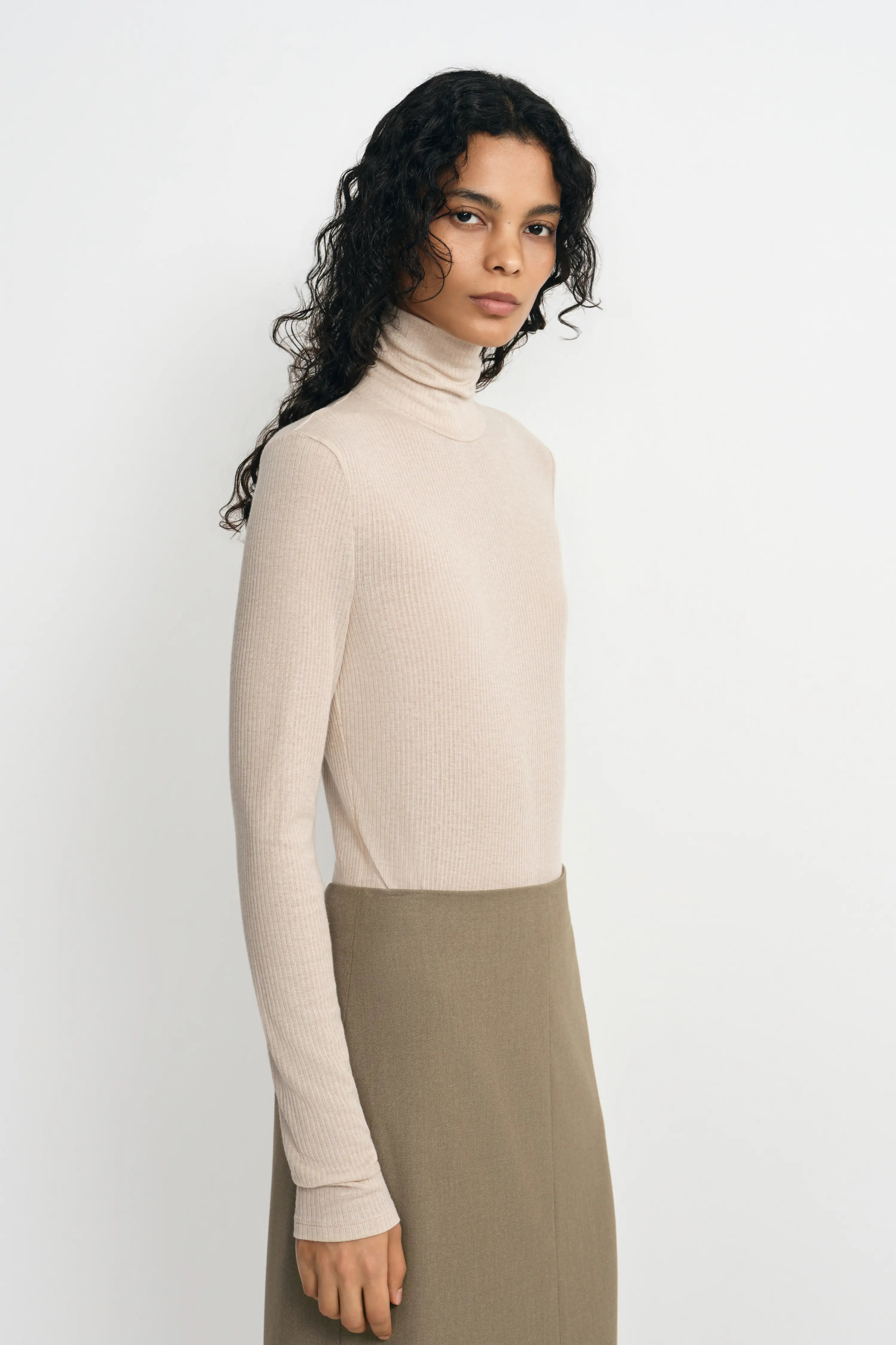 A woman modeling a ribbed long-sleeve turtleneck sweater in a light beige color, paired with a taupe skirt, against a plain light gray studio background.