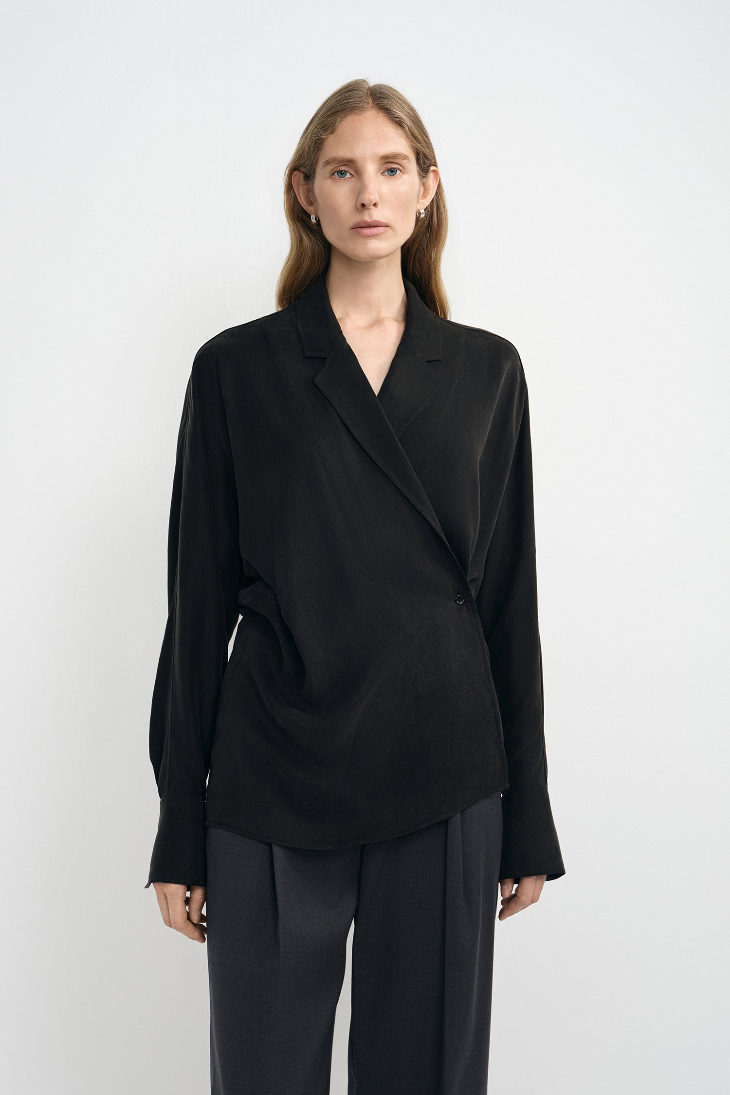 A woman modeling a black wrap-style long-sleeve blouse with draped detailing and dark wide-leg trousers, photographed against a clean light gray studio background.