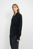 A woman modeling a black long-sleeve wrap-style blouse with a twisted waist detail, paired with dark trousers, against a plain light gray studio background.
