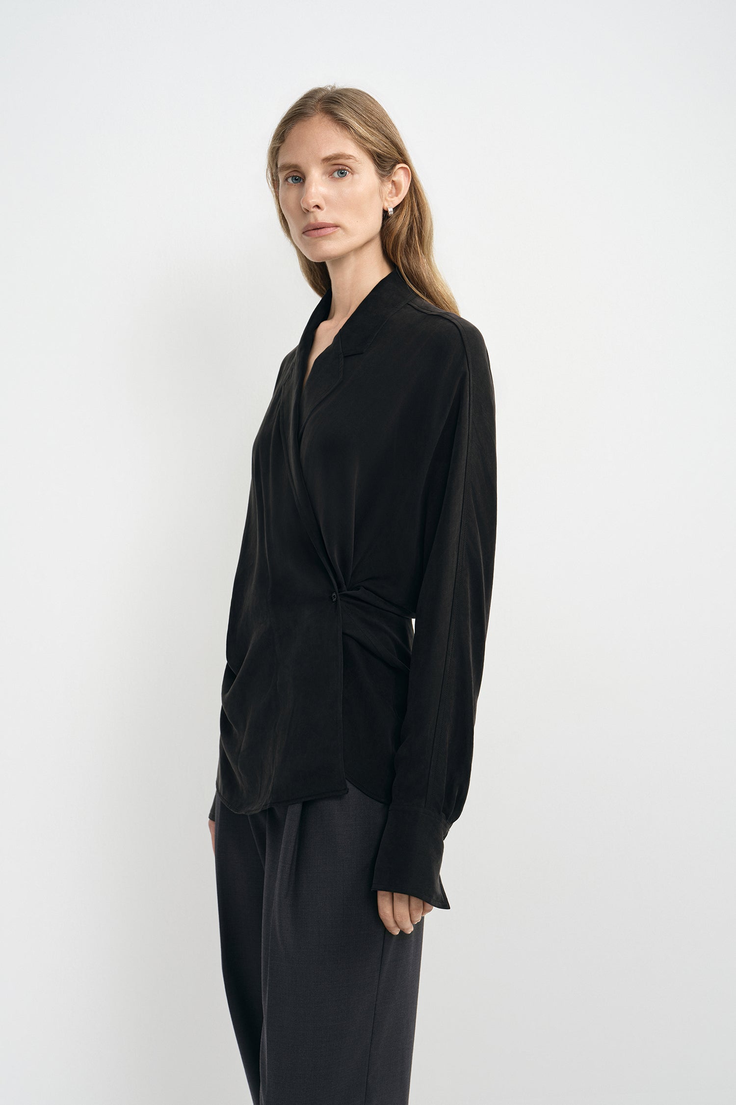 A woman modeling a black long-sleeve wrap-style blouse with a twisted waist detail, paired with dark trousers, against a plain light gray studio background.