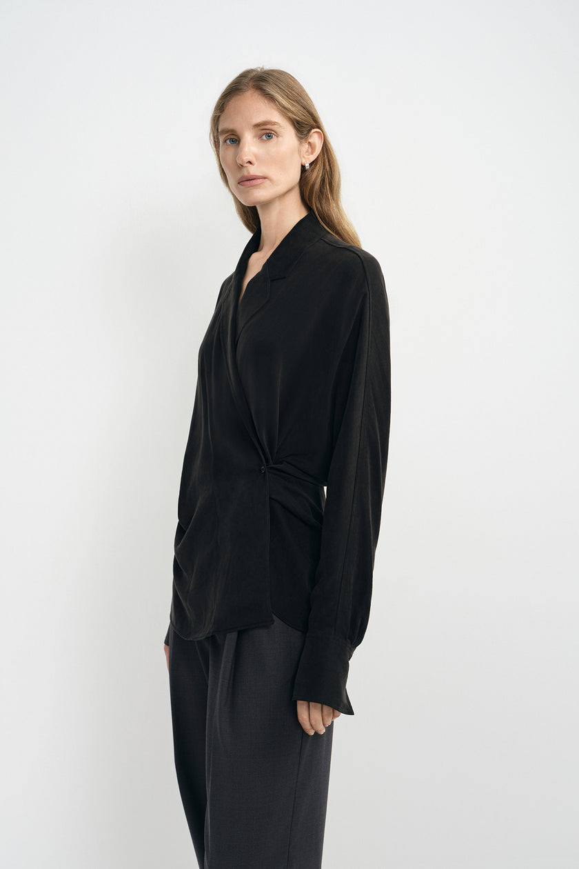 A woman modeling a black long-sleeve wrap-style blouse with a twisted waist detail, paired with dark trousers, against a plain light gray studio background.