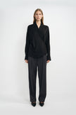 A model wearing dark grey pleat-front wide-leg trousers in a silky wool blend, pairing an oversized black wrap-front blouse and black pointed-toe heels.