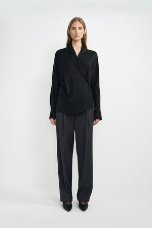 A model wearing dark grey pleat-front wide-leg trousers in a silky wool blend, pairing an oversized black wrap-front blouse and black pointed-toe heels.