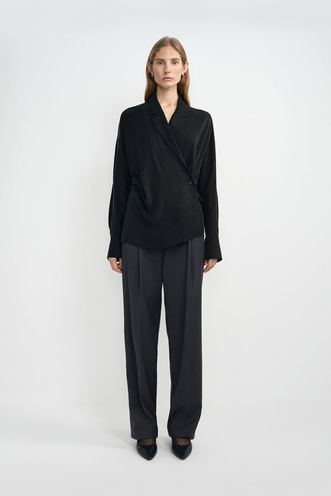 A model wearing dark grey pleat-front wide-leg trousers in a silky wool blend, pairing an oversized black wrap-front blouse and black pointed-toe heels.