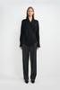 A model wearing dark grey pleat-front wide-leg trousers in a silky wool blend, pairing an oversized black wrap-front blouse and black pointed-toe heels.