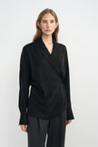 A woman modeling a black long-sleeve wrap-style blouse with a relaxed, draped silhouette and dark trousers, photographed against a clean light gray studio background.