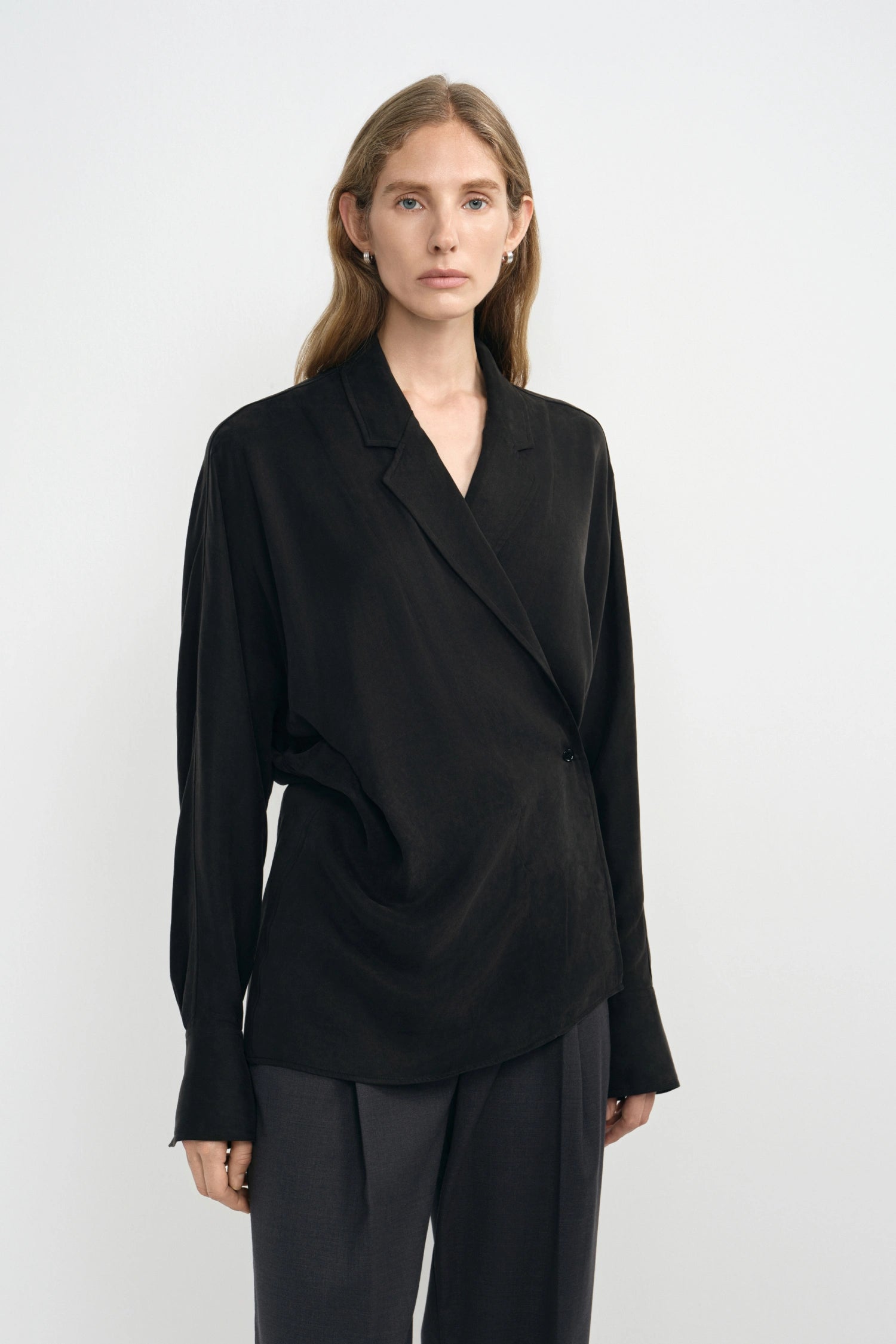 A woman modeling a black long-sleeve wrap-style blouse with a relaxed, draped silhouette and dark trousers, photographed against a clean light gray studio background.