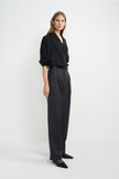 A woman modeling a black wrap-style blouse with rolled sleeves and high-waisted dark charcoal wide-leg trousers, standing against a clean light gray studio background.