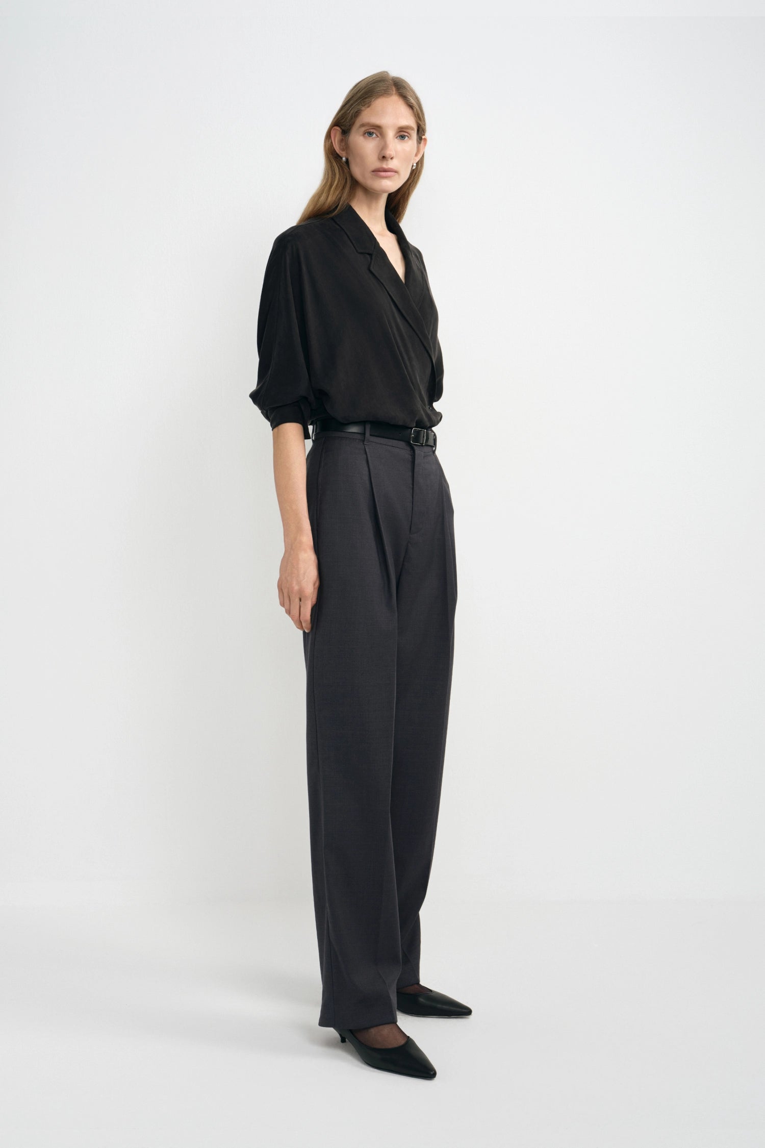 A woman modeling a black wrap-style blouse with rolled sleeves and high-waisted dark charcoal wide-leg trousers, standing against a clean light gray studio background.