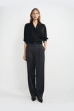 A model wearing dark grey pleat-front wide-leg trousers in a silky wool blend, pairing a black wrap-front blouse and black pointed-toe heels.
