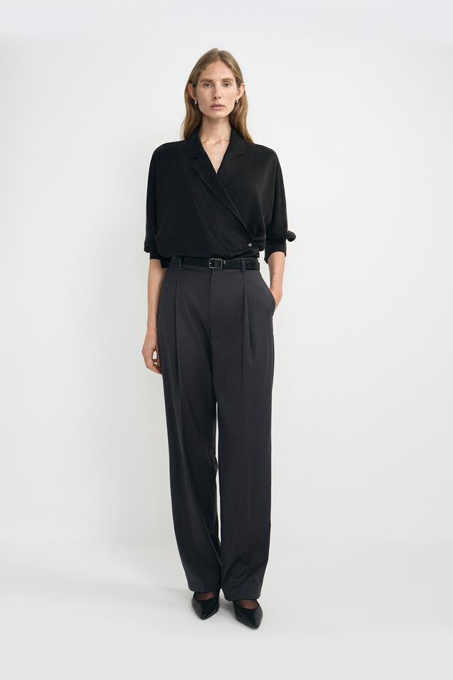 A model wearing dark grey pleat-front wide-leg trousers in a silky wool blend, pairing a black wrap-front blouse and black pointed-toe heels.