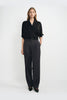 A model wearing dark grey pleat-front wide-leg trousers in a silky wool blend, pairing a black wrap-front blouse and black pointed-toe heels.