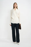 A model stands against a plain white studio background wearing a cream-colored wrap-style blouse with dark wide-leg trousers and white pointed shoes, holding a brown clutch.