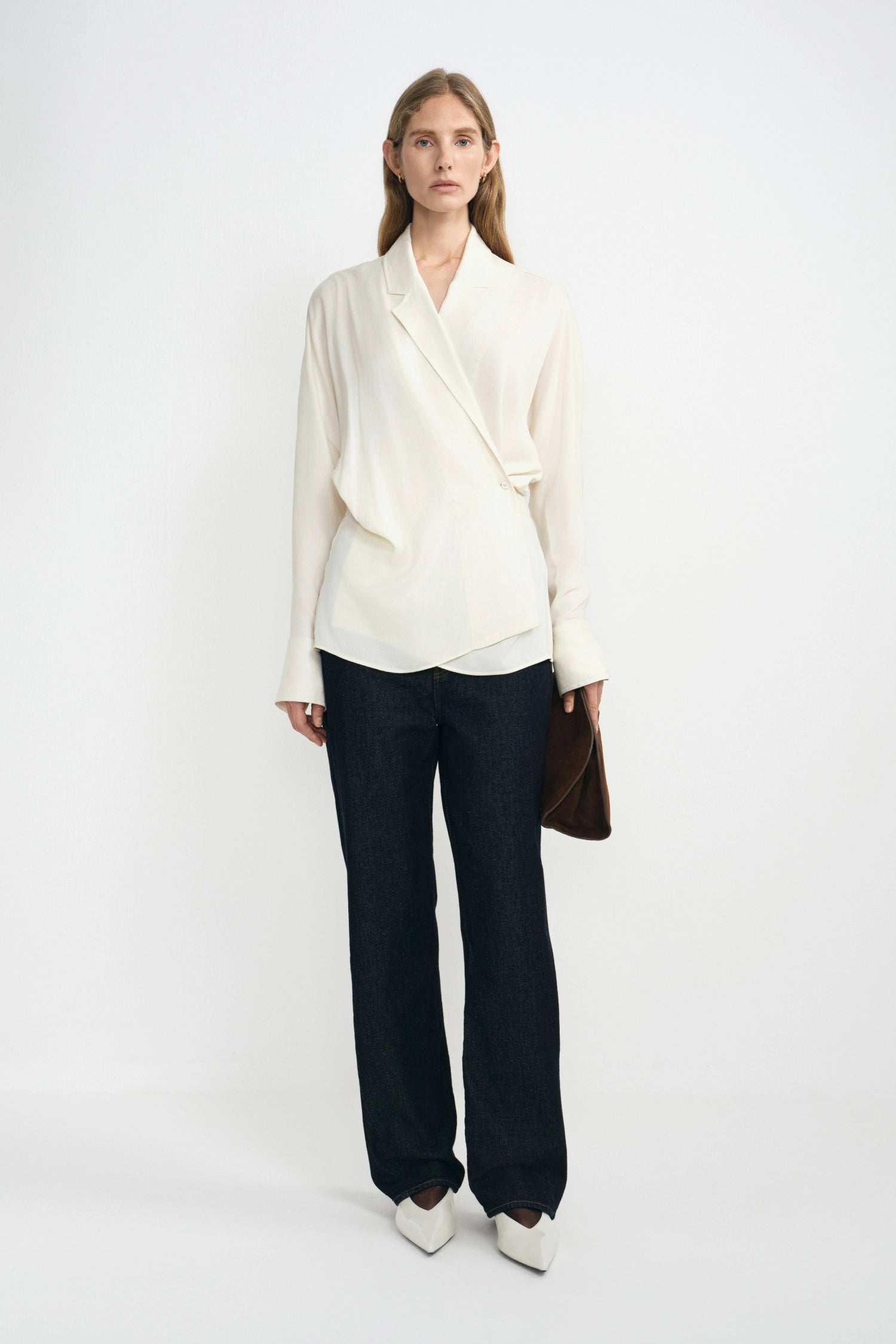 A model stands against a plain white studio background wearing a cream-colored wrap-style blouse with dark wide-leg trousers and white pointed shoes, holding a brown clutch.