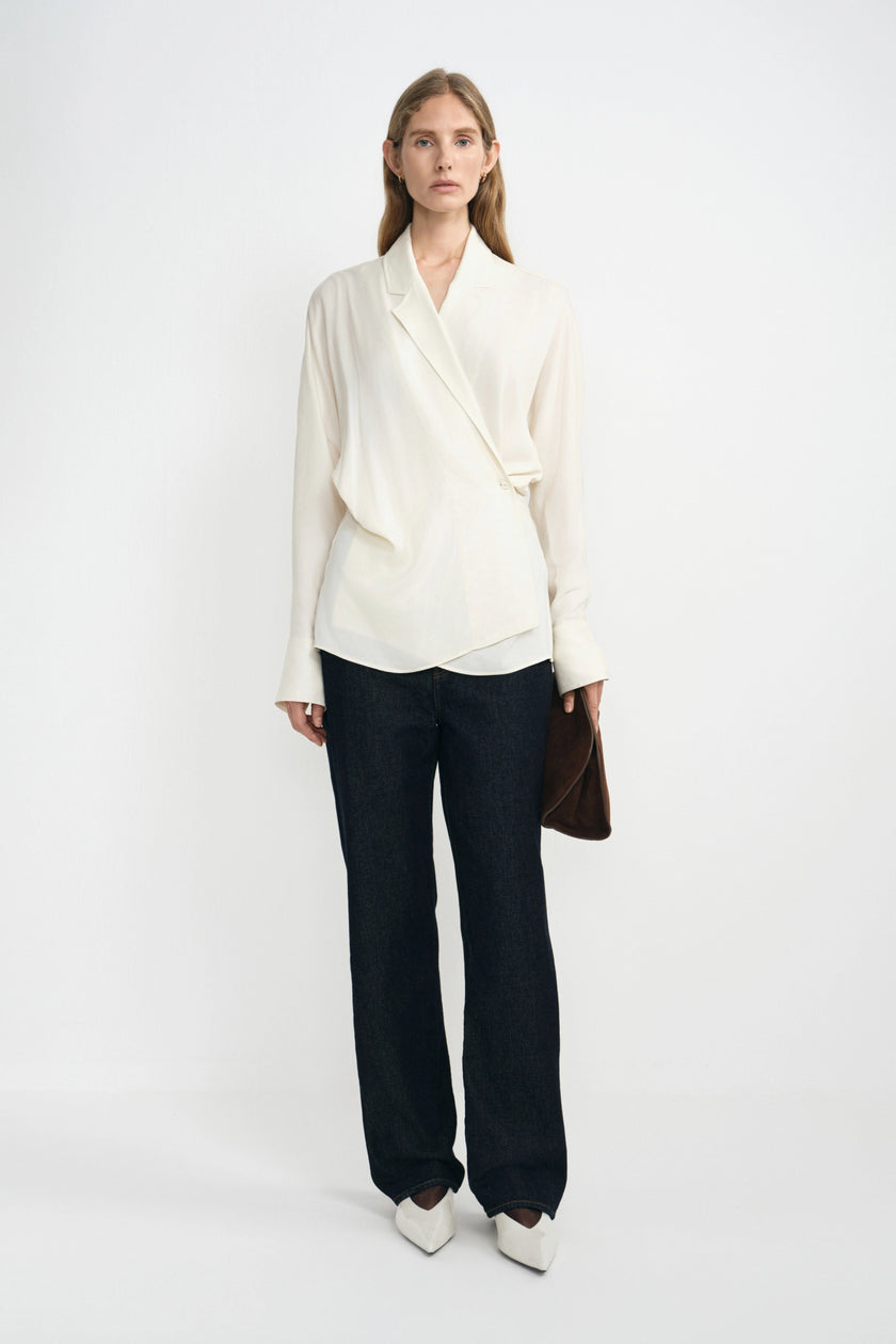 A model stands against a plain white studio background wearing a cream-colored wrap-style blouse with dark wide-leg trousers and white pointed shoes, holding a brown clutch.
