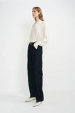 A model stands against a plain white studio background wearing a cream-colored wrap-style blouse with dark wide-leg jeans, a black belt, and white pointed-toe shoes.