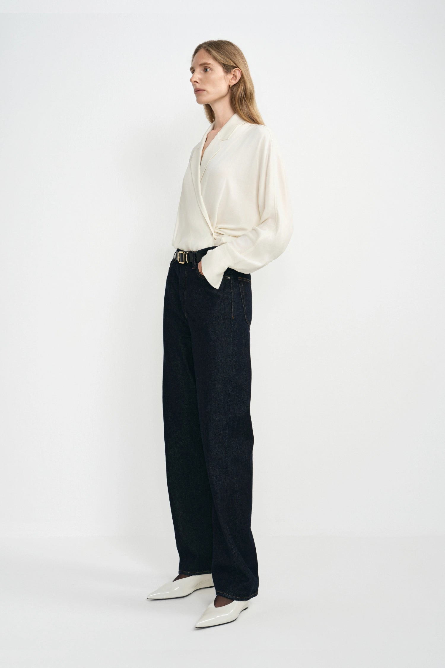 A model stands against a plain white studio background wearing a cream-colored wrap-style blouse with dark wide-leg jeans, a black belt, and white pointed-toe shoes.