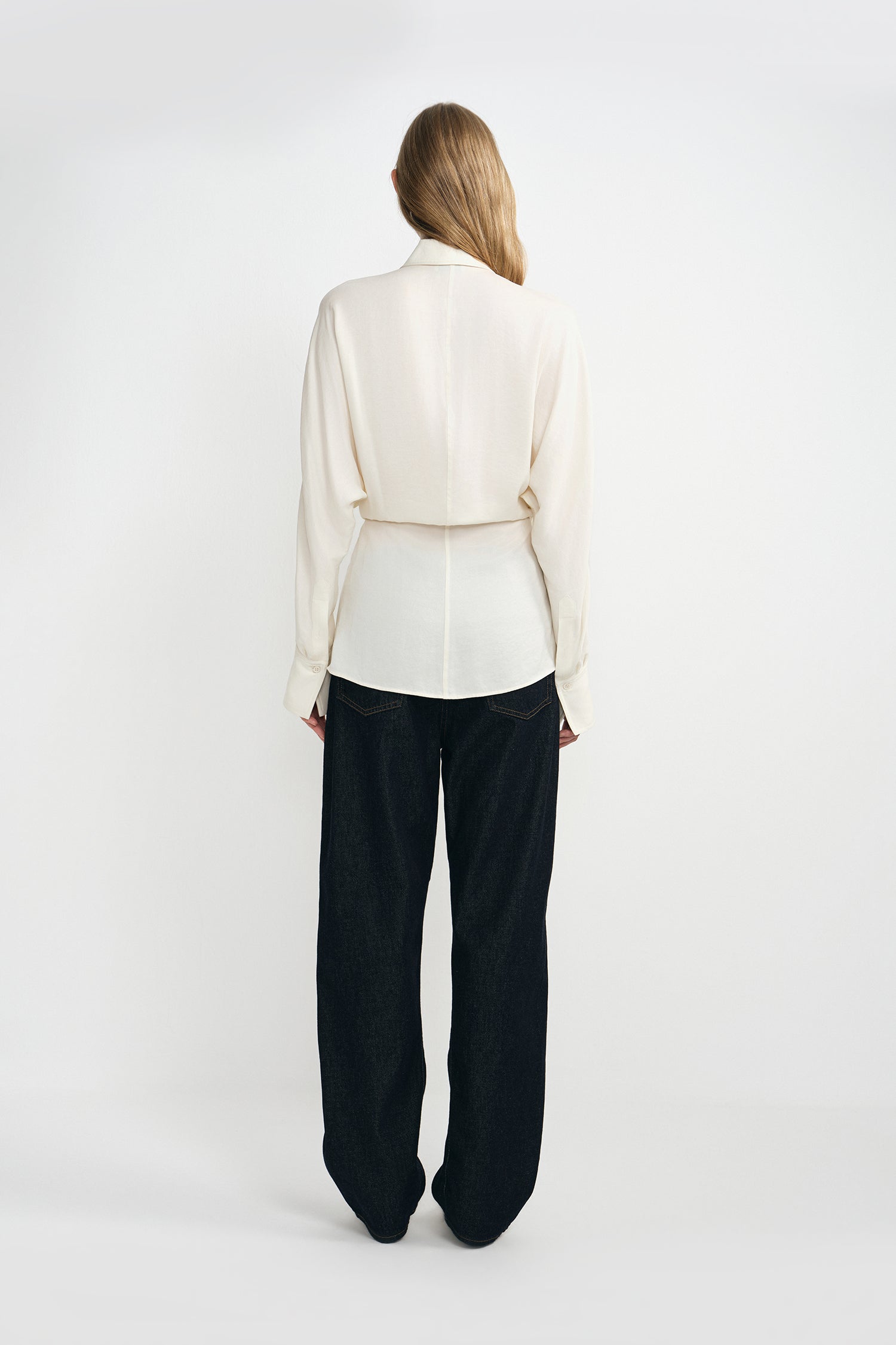 A woman modeling a cream-colored silky wrap-style shirt with long sleeves and classic dark blue jeans.