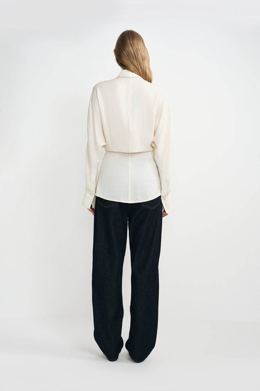 A woman modeling a cream-colored silky wrap-style shirt with long sleeves and classic dark blue jeans.