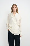 A woman modeling a cream-colored silky wrap-style shirt with long sleeves and classic dark blue jeans.
