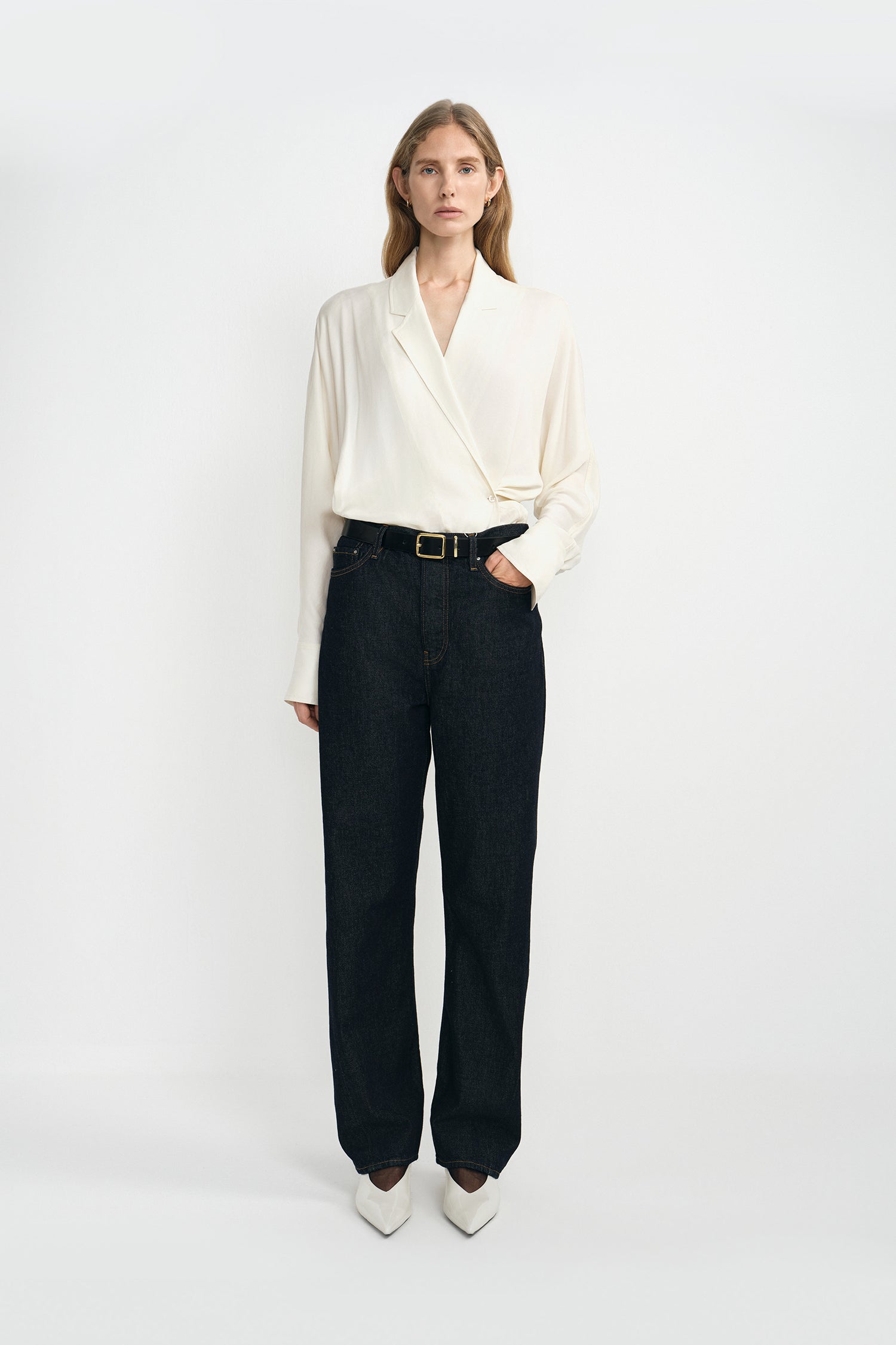 A woman modeling a cream-colored silky wrap-style shirt with long sleeves and classic dark blue jeans, with a classic black cow leather belt, and white pointed kitten heels.