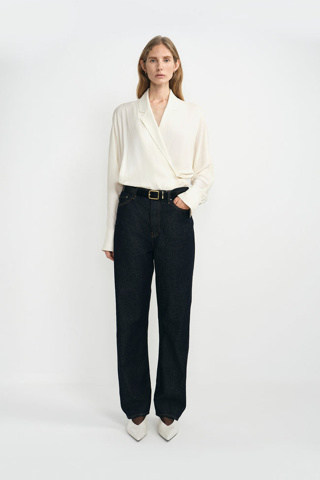 A woman modeling a cream-colored silky wrap-style shirt with long sleeves and classic dark blue jeans, with a classic black cow leather belt, and white pointed kitten heels.