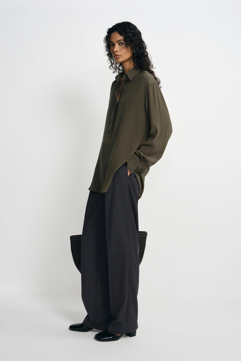 A model stands in profile wearing an oversized olive green button-up shirt and very wide-leg dark charcoal trousers, photographed against a clean light gray studio background.