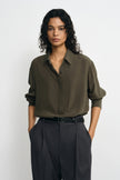 A woman modeling a long-sleeve olive green button-up shirt tucked into dark charcoal pleated trousers, standing against a light gray studio background.