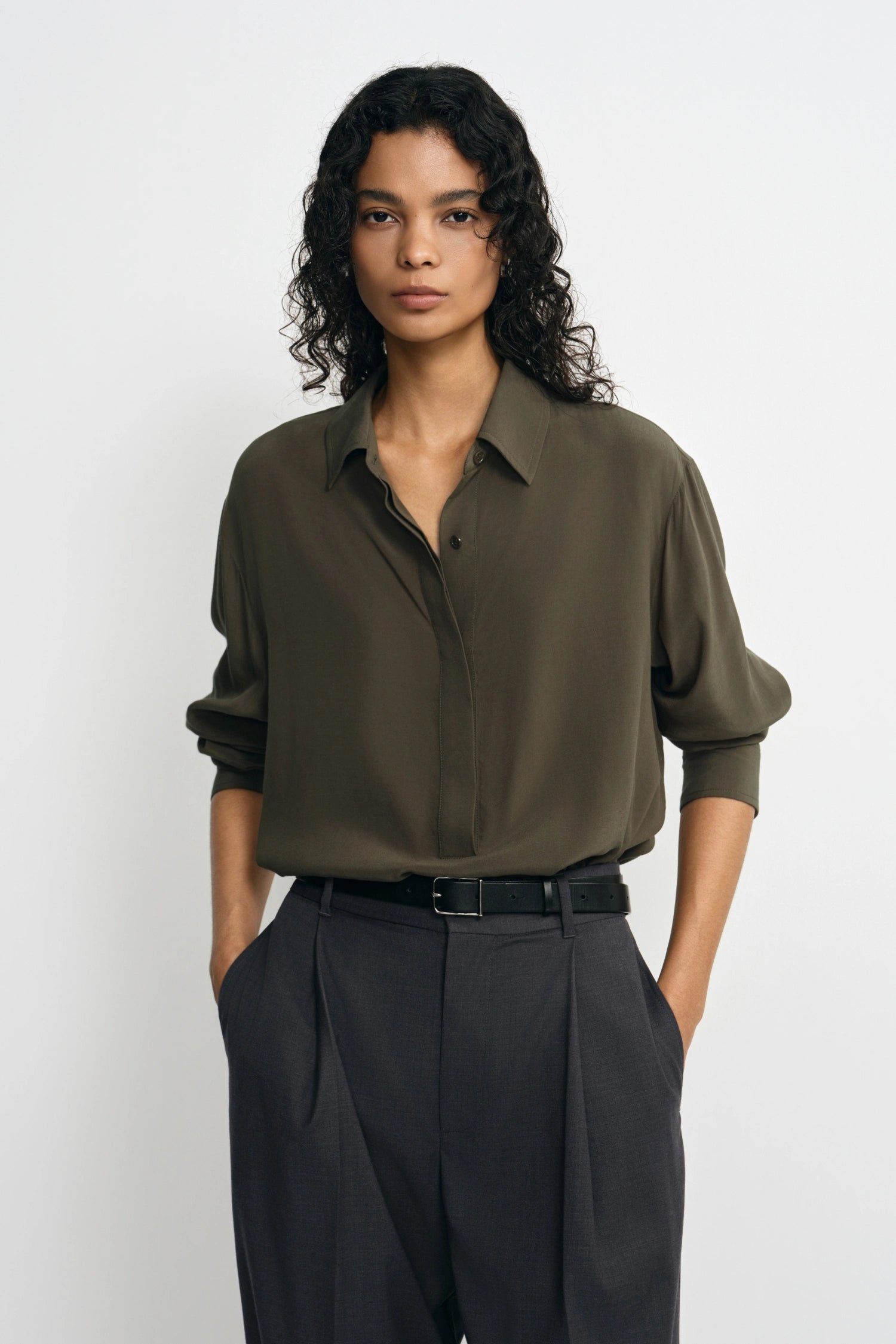 A woman modeling a long-sleeve olive green button-up shirt tucked into dark charcoal pleated trousers, standing against a light gray studio background.