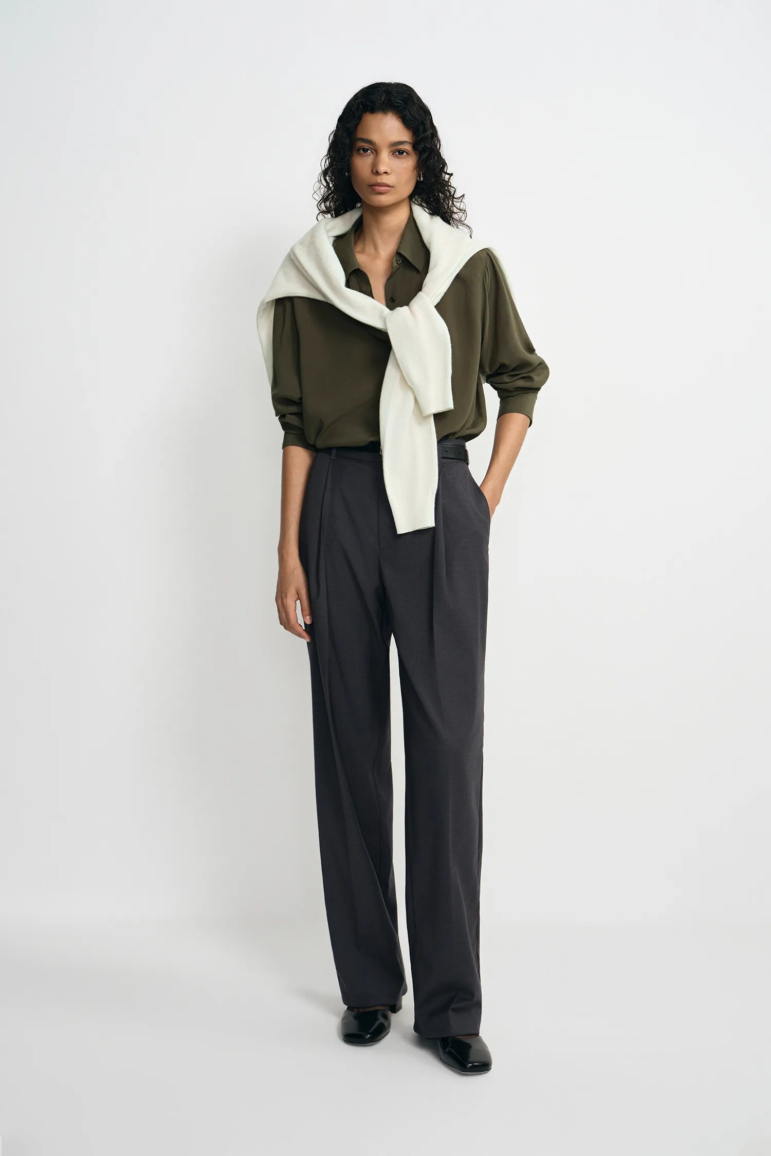 A model stands against a light gray studio background wearing a dark olive green button-up shirt, dark charcoal gray wide-leg trousers, and a cream-colored knit wrap tied around her shoulders.