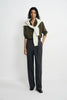 A model stands against a light gray studio background wearing a dark olive green button-up shirt, dark charcoal gray wide-leg trousers, and a cream-colored knit wrap tied around her shoulders.