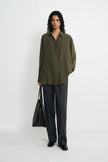 A model stands against a plain white studio background wearing an oversized olive green button-up shirt, dark charcoal gray wide-leg trousers, and black shoes, while holding a black tote bag.