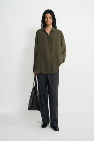 A model stands against a plain white studio background wearing an oversized olive green button-up shirt, dark charcoal gray wide-leg trousers, and black shoes, while holding a black tote bag.
