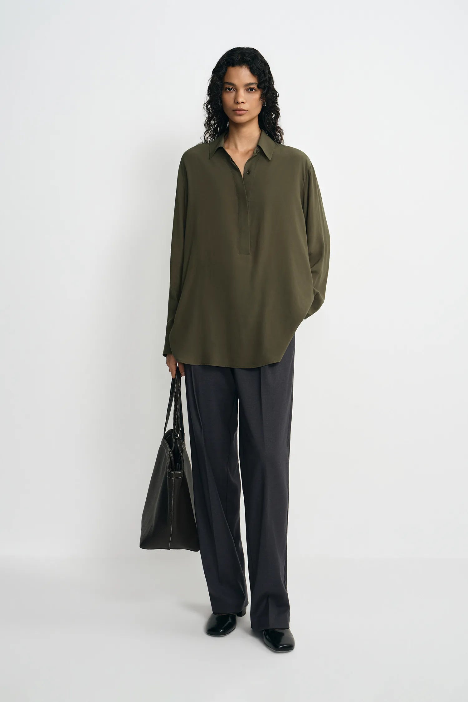 A model stands against a plain white studio background wearing an oversized olive green button-up shirt, dark charcoal gray wide-leg trousers, and black shoes, while holding a black tote bag.