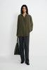 A model stands against a plain white studio background wearing an oversized olive green button-up shirt, dark charcoal gray wide-leg trousers, and black shoes, while holding a black tote bag.