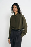 A model wearing a dark olive green long-sleeve button-up shirt tucked into dark charcoal gray pleated trousers, standing against a plain light gray studio background.