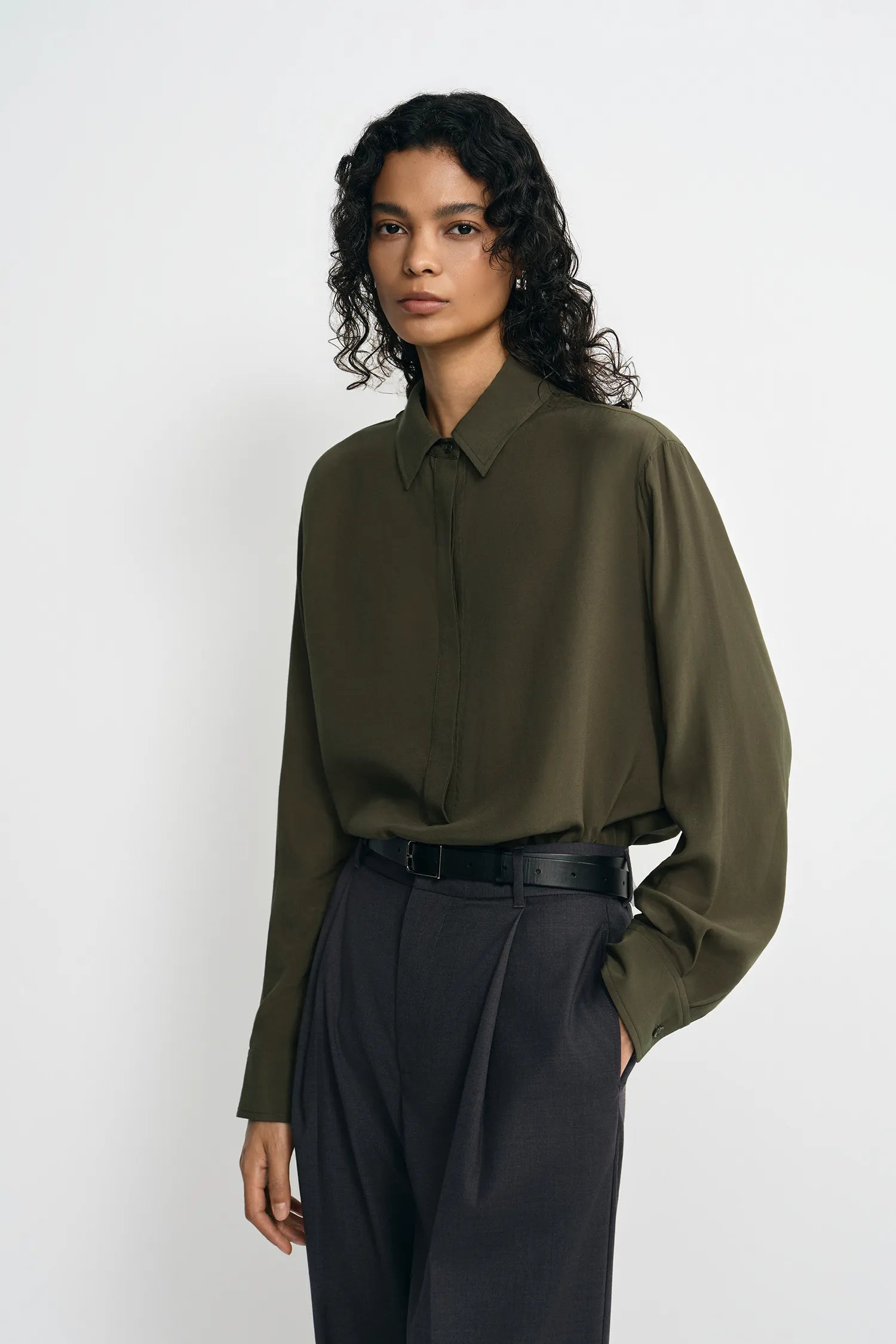 A model wearing a dark olive green long-sleeve button-up shirt tucked into dark charcoal gray pleated trousers, standing against a plain light gray studio background.