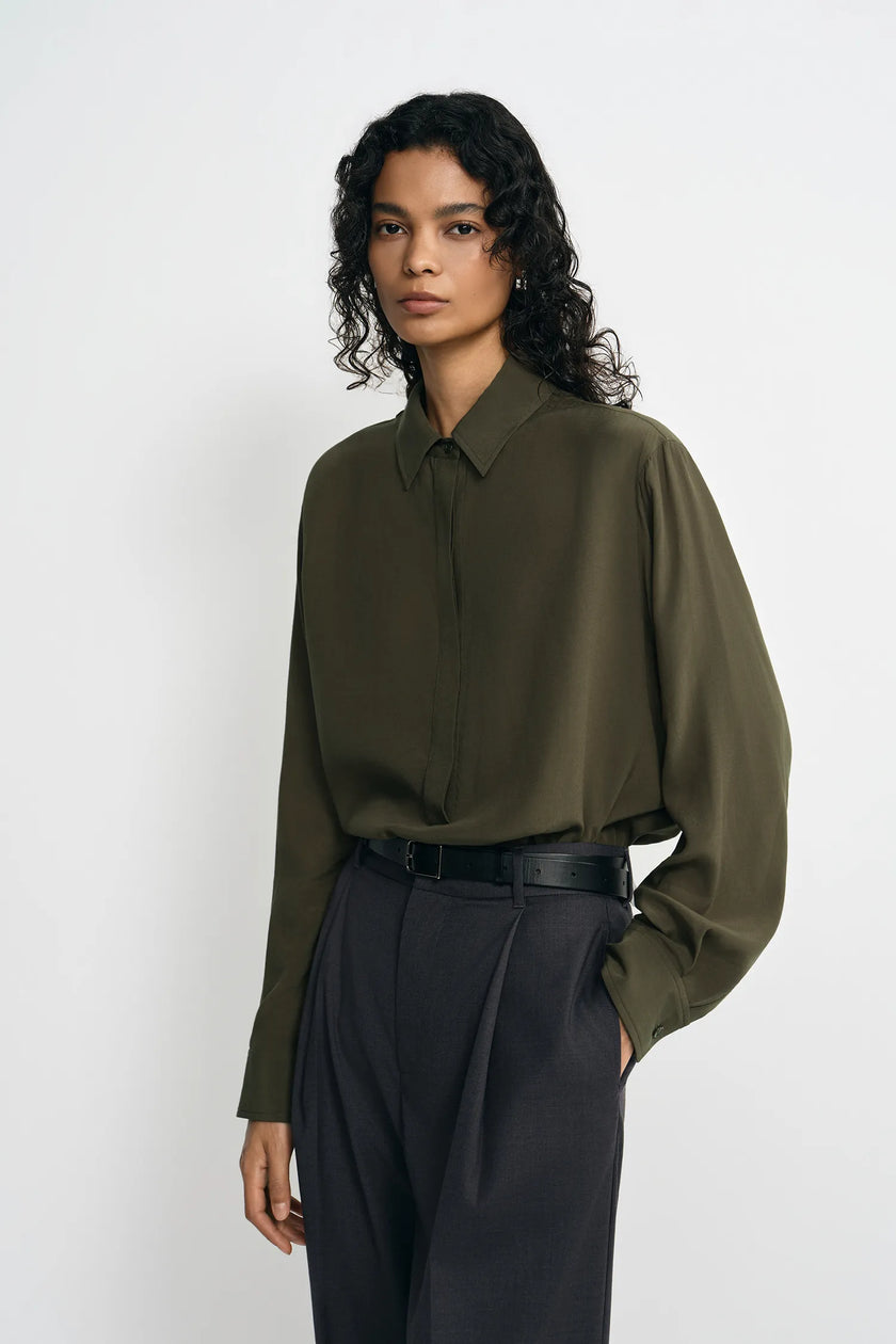 A model wearing a dark olive green long-sleeve button-up shirt tucked into dark charcoal gray pleated trousers, standing against a plain light gray studio background.