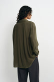 A woman shown from the back wearing an oversized olive green button-up shirt and dark charcoal pants, standing against a plain light gray studio background.