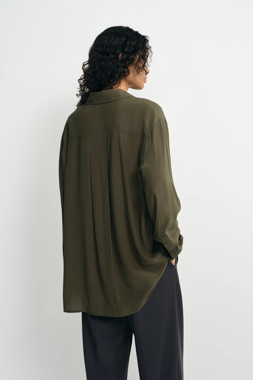 A woman shown from the back wearing an oversized olive green button-up shirt and dark charcoal pants, standing against a plain light gray studio background.