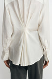 Close-up of a winter-white blouse on a model from behind, showing pleats drawn together at the waist and fanning out toward the curved hem.